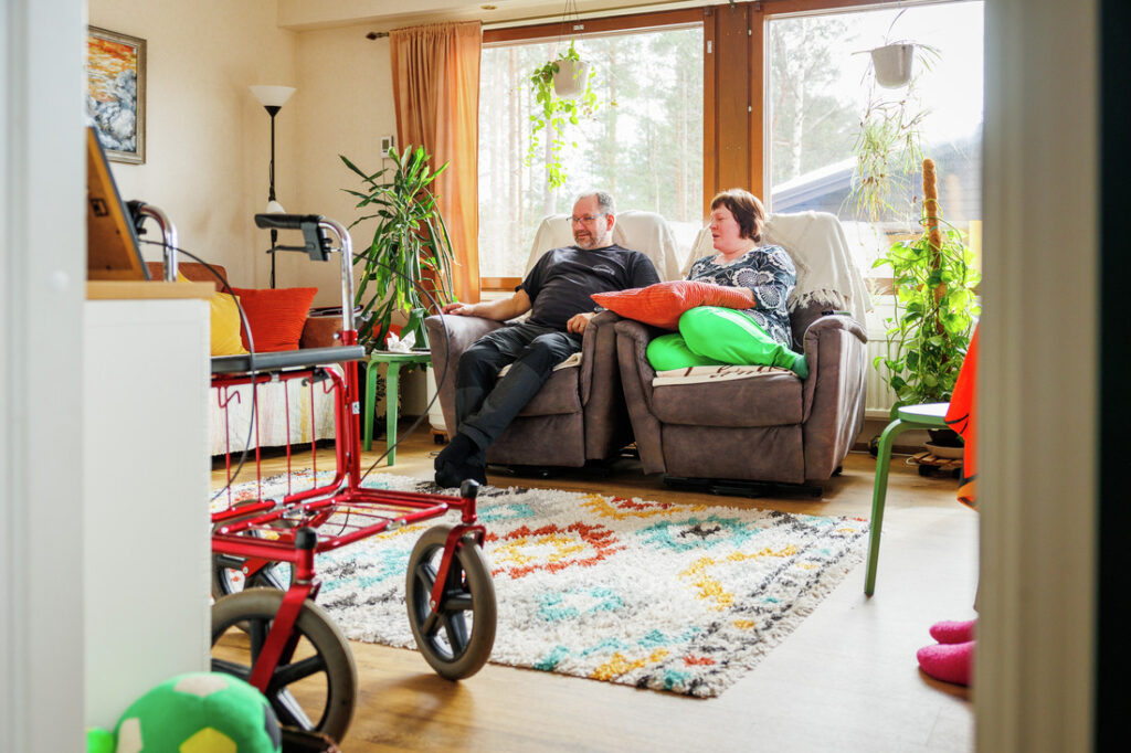 Perhehoito paikan pitäjät Timo Tuononen (vasemmalla) ja Taru Tuononen ( oikealla) istuvat tuolleissa olohuoneessa. Edustalla on punainen rollaattori.