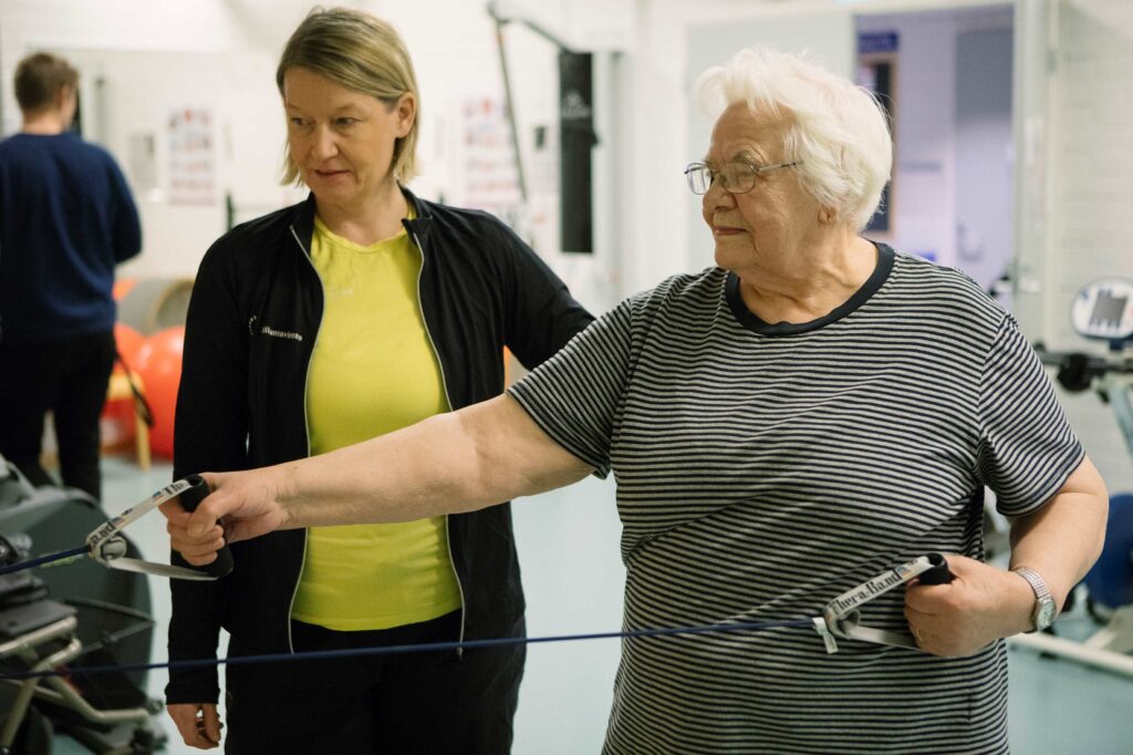 Ikääntynyt henkilö jumppaa, ohjaaja seuraa vieressä.