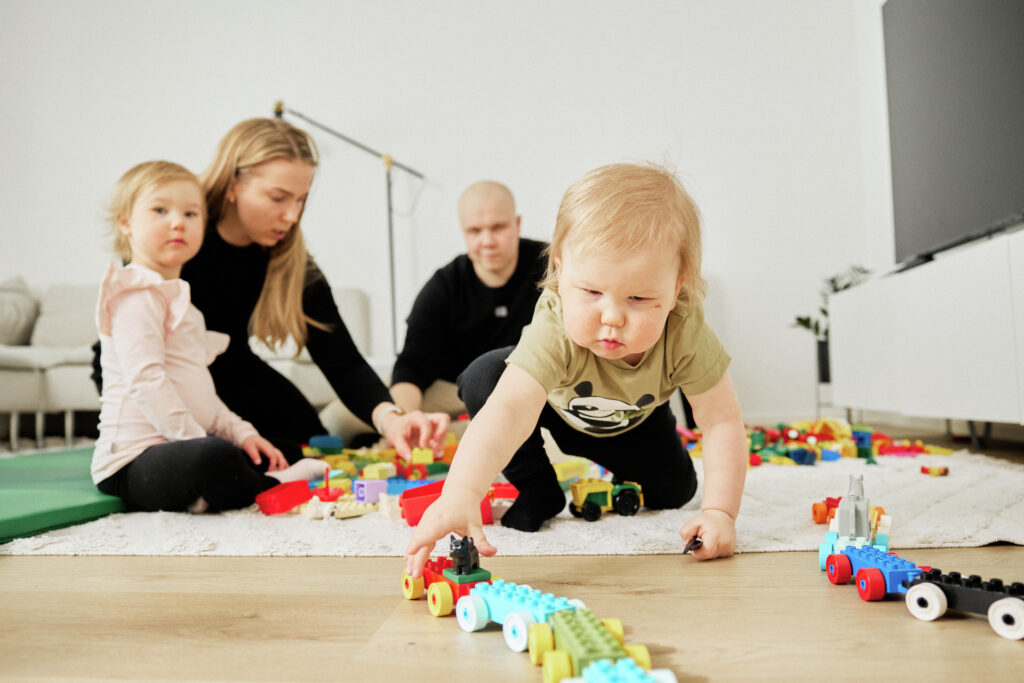 Lapsi etualalla vetää leikki junaa. Taustalla vanhemmat rakentavat dubloista rakennuksia vanhemman lapsen kanssa.