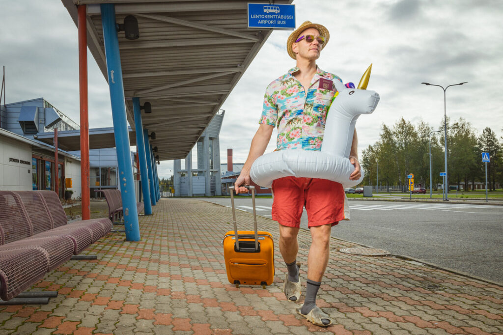 Lomailija kävelemässä joensuun lentoasemalla vetäen perässään matkalaukkua. Päällä havaiji-paita ja yksisarvinen uimalelu