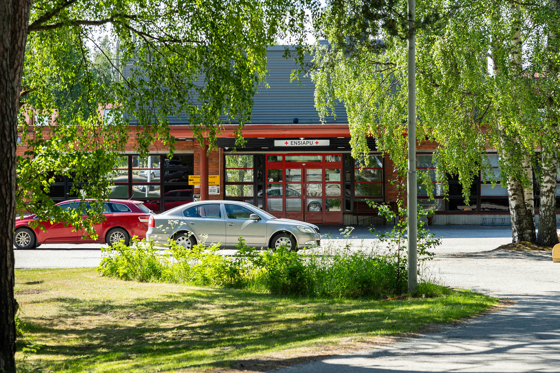 Nurmeksen sosiaali- ja terveyskeskuksen rakennus ulkoapäin, etualalla kaksi henkilöautoa ja kesäisiä koivujen oksia